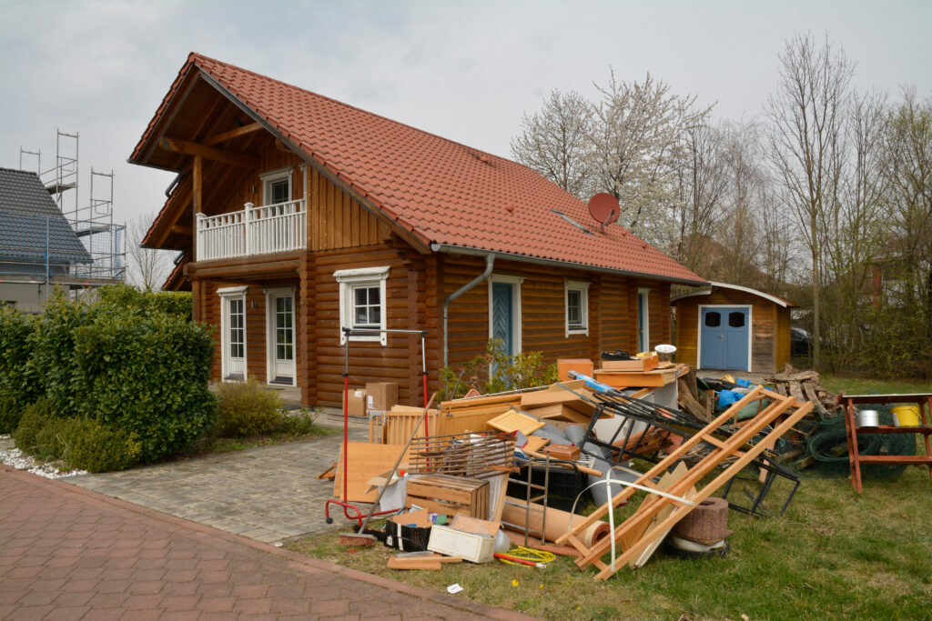 Ein Holzhaus im ländlichen Bereich mit einem Haufen von Möbeln und Bauabfällen vor dem Haus. Der Garten ist unordentlich, und es gibt verschiedene Gegenstände wie Holzstücke, Kisten und Möbelteile, die auf dem Rasen gestapelt sind.